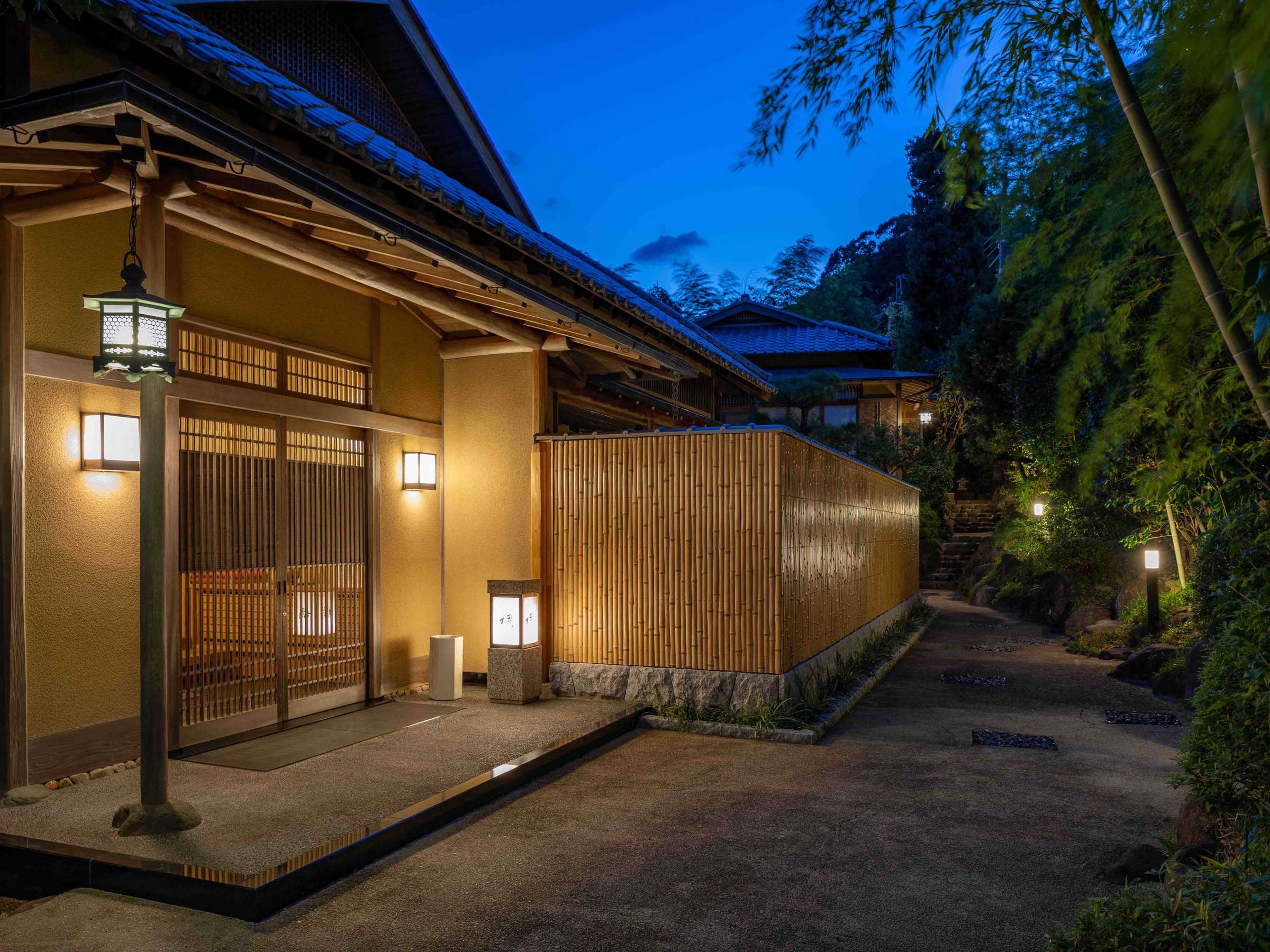 Lantern-lit main entrance approach with bamboo fence and stone path at dusk at Atami Koarashitei ryokan