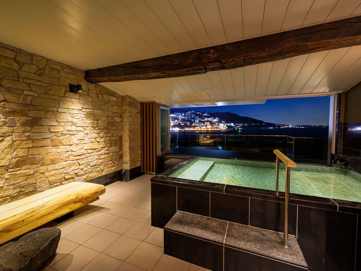 Stone-walled open-air rotenburo bath at night with Atami city lights across the bay