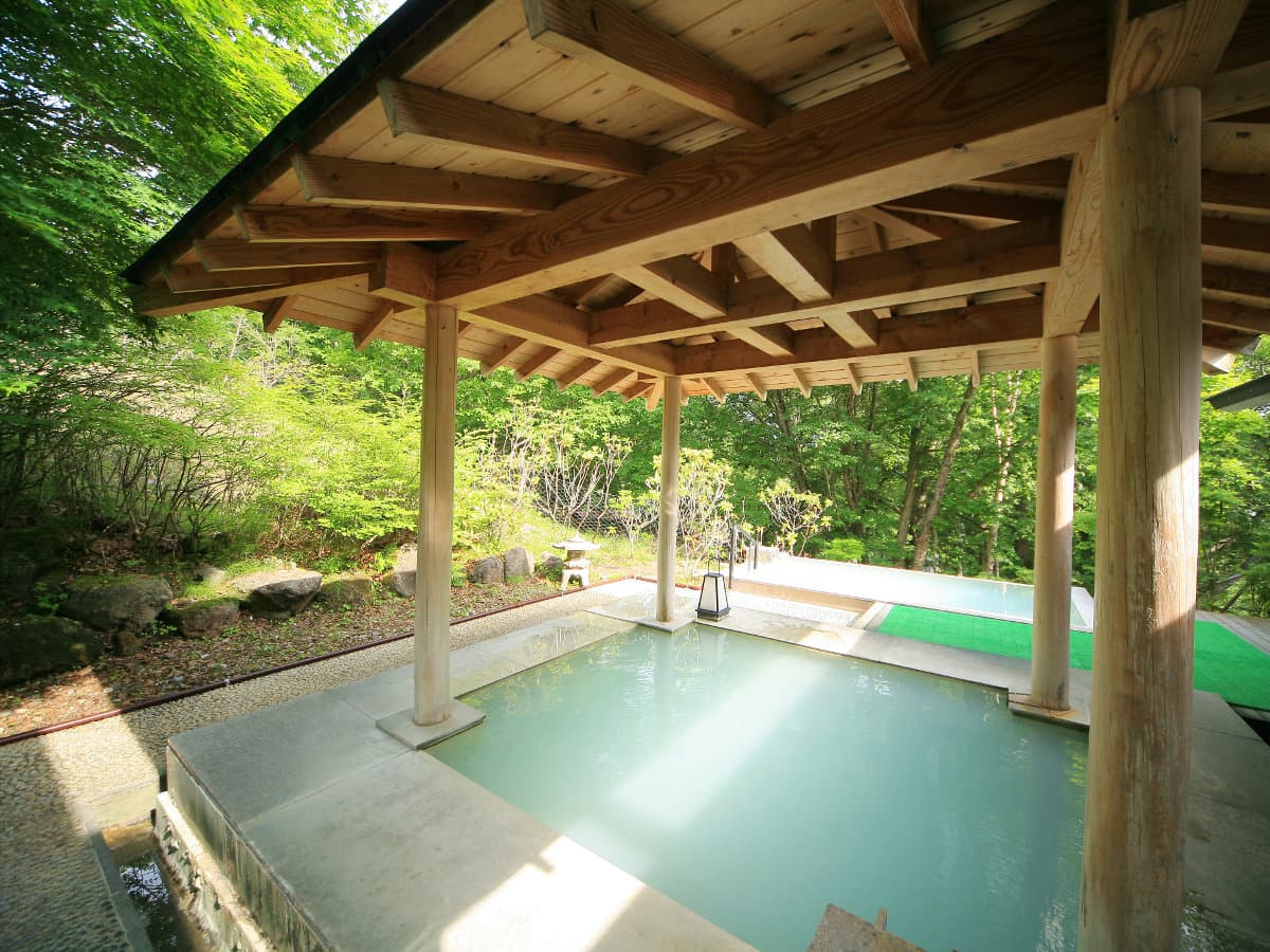 Open-air stone rotenburo onsen bath under a wooden pavilion surrounded by green forest at Okunikko Hotel Shikisai