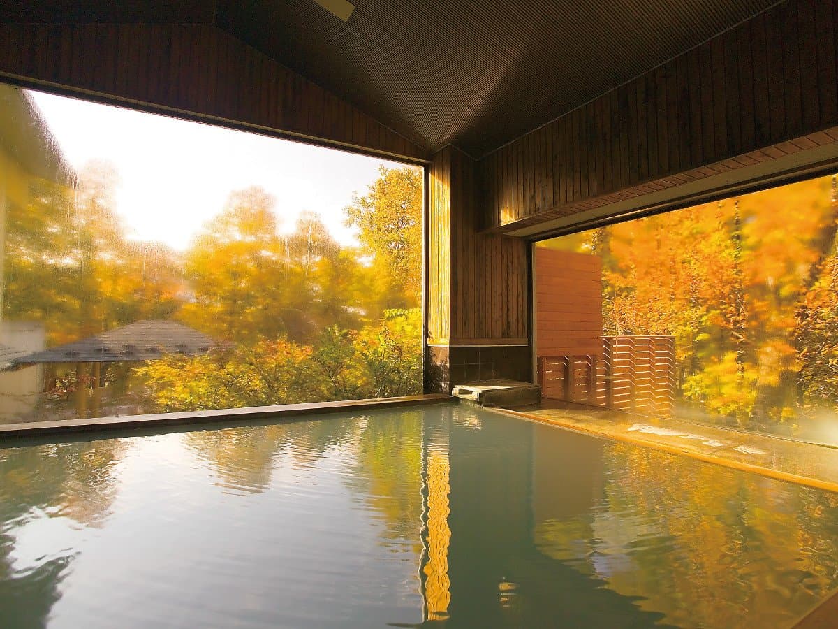 Indoor onsen bath with large windows framing golden autumn foliage at Okunikko Hotel Shikisai