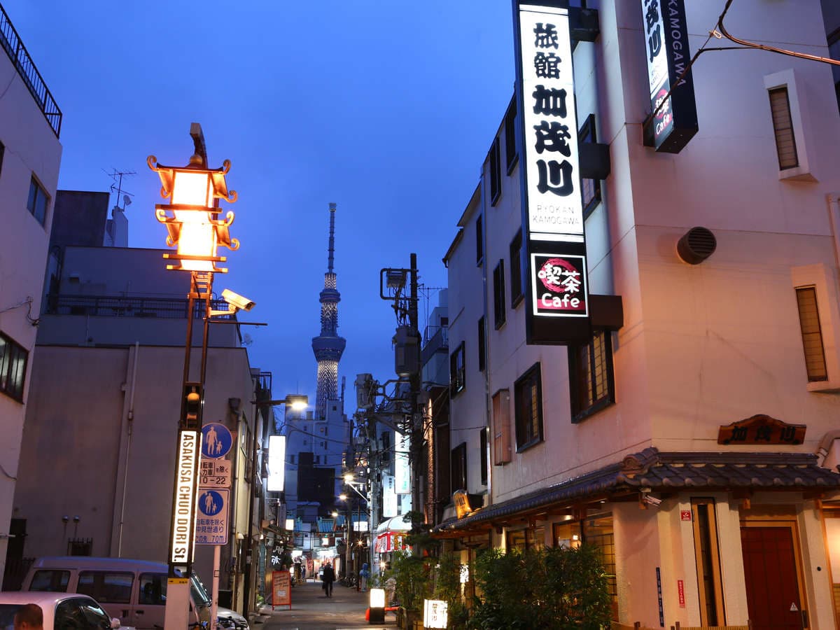 Ryokan Kamogawa exterior at dusk with its illuminated sign on a quiet Asakusa lane, Tokyo Skytree rising in the distance