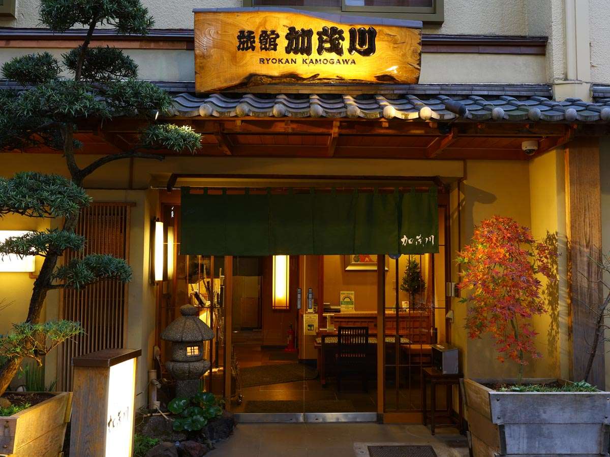 Warmly lit traditional entrance of Ryokan Kamogawa in Asakusa, Tokyo, with green noren curtain, wooden sign, stone lantern and autumn maple