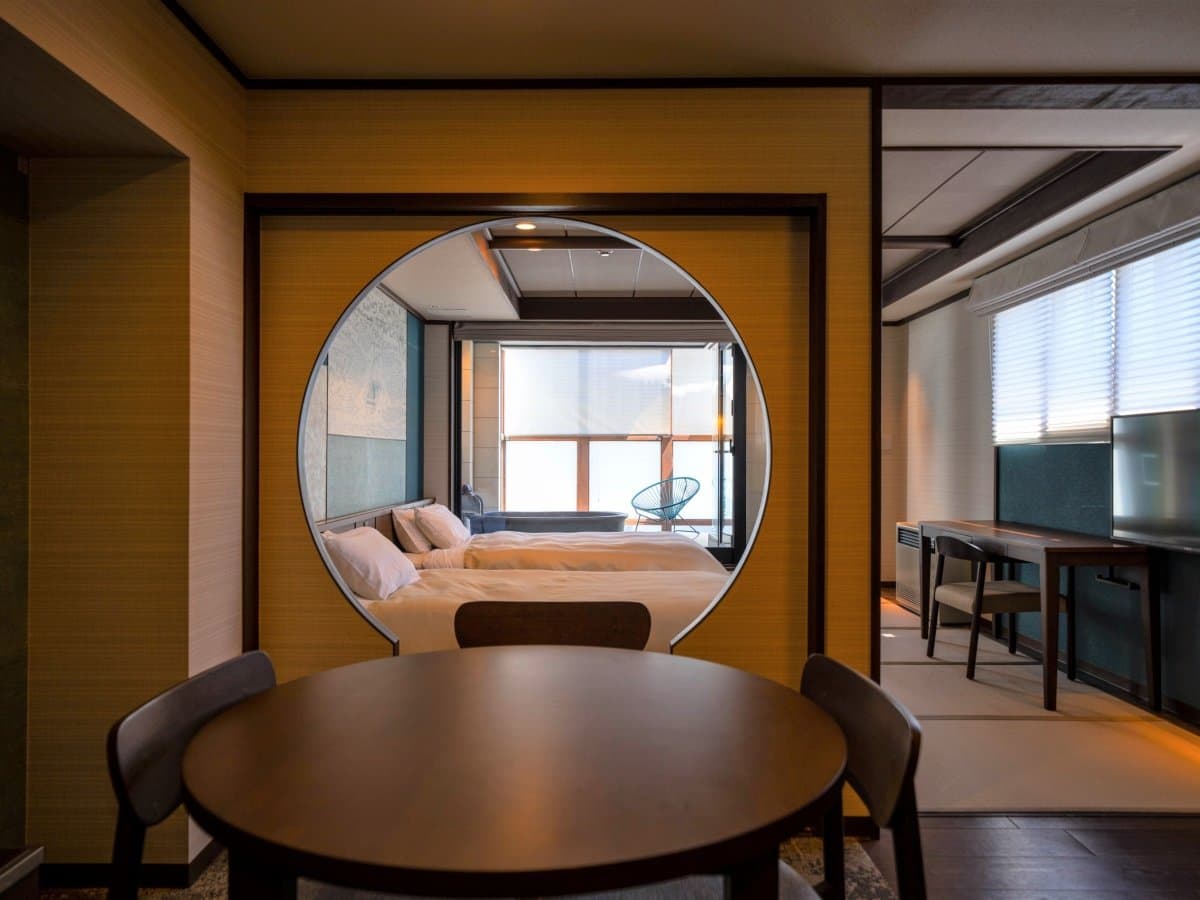 Modern guest room viewed through a round moon-window framing twin beds and a dining table