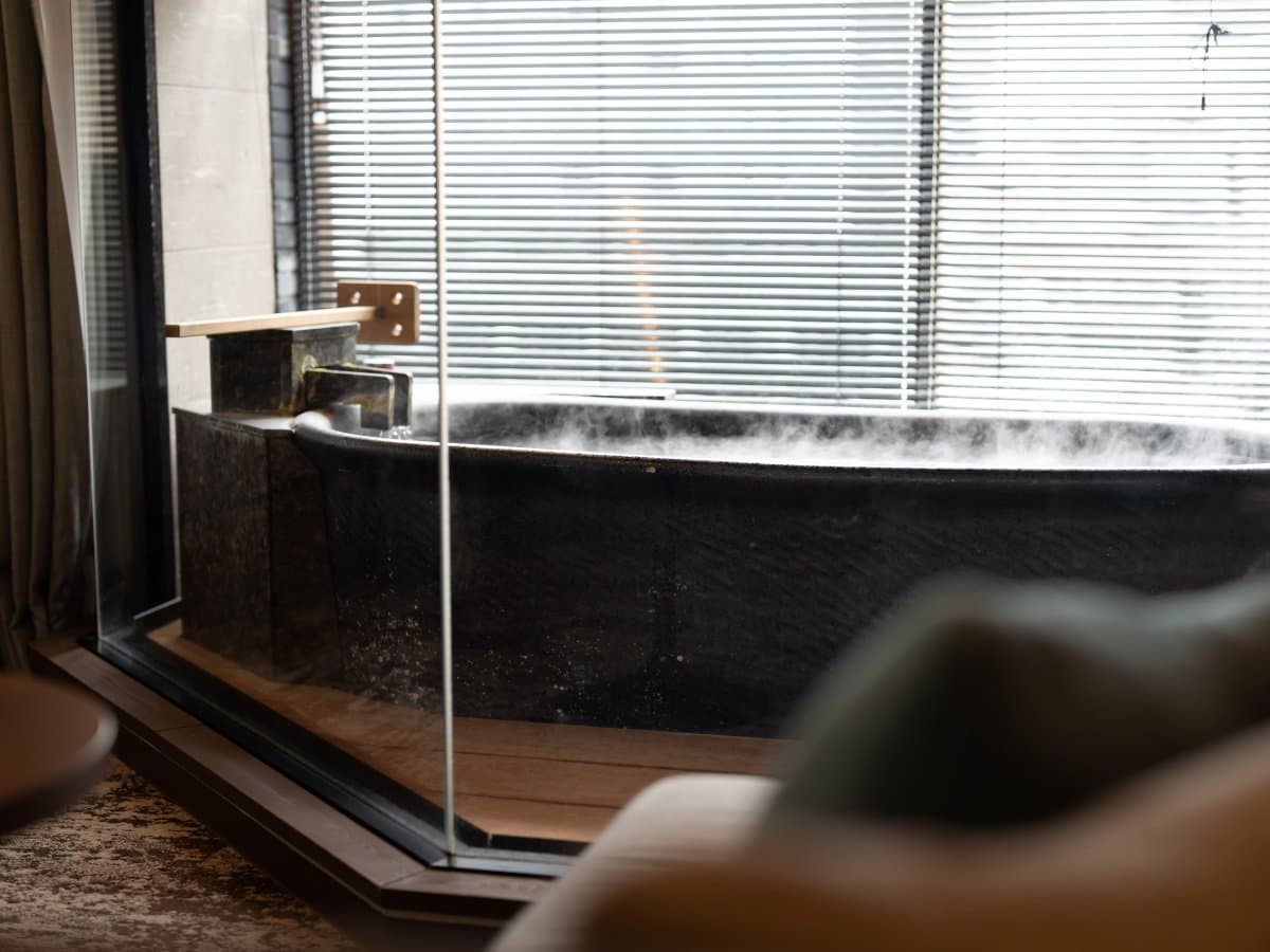 In-room private soaking tub steaming beside a bright shaded window in a guest room