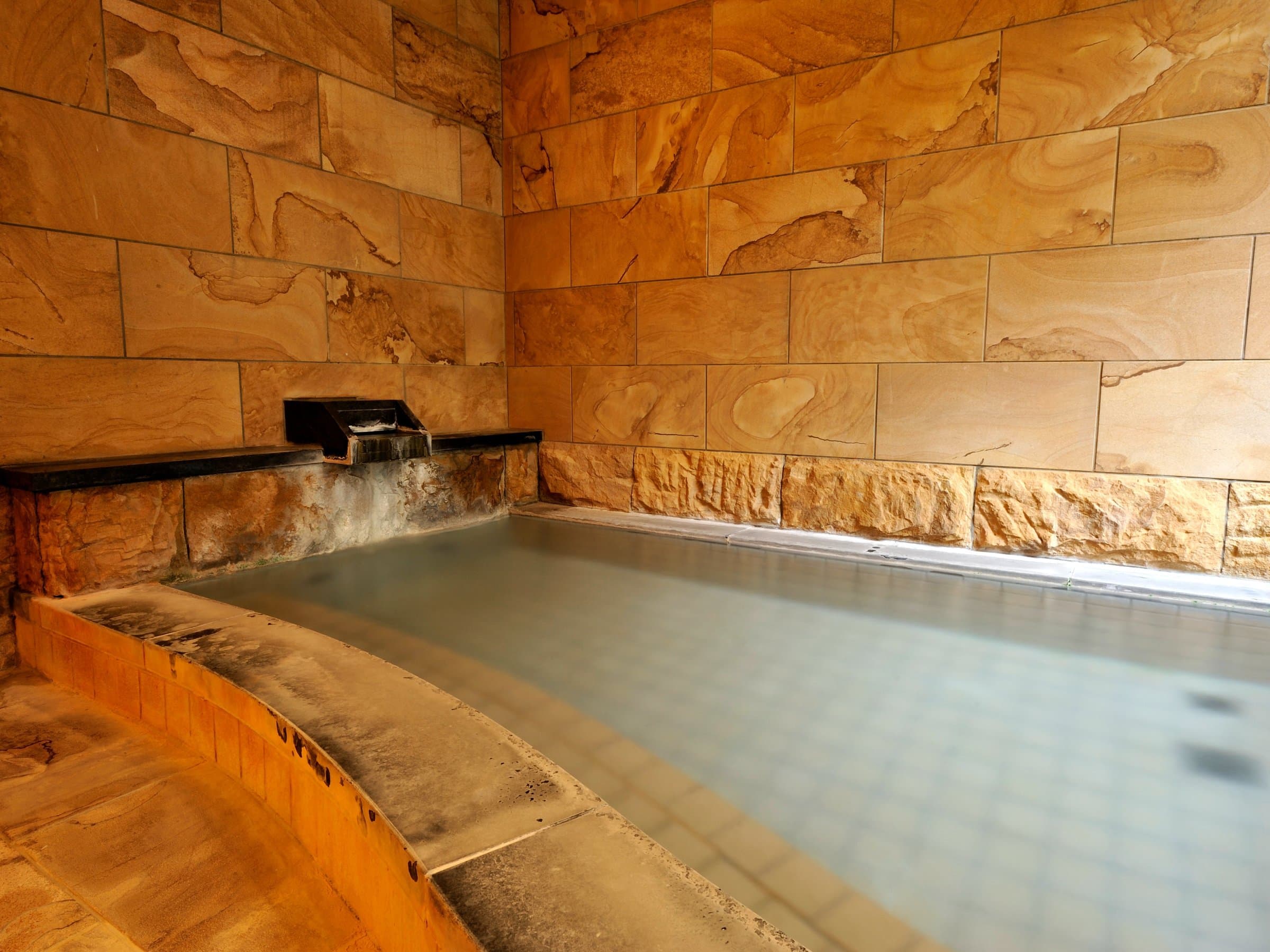 Steaming indoor hot spring tub clad in warm sandstone with a stone spout at Shirouma-so, Hakuba Happo Onsen