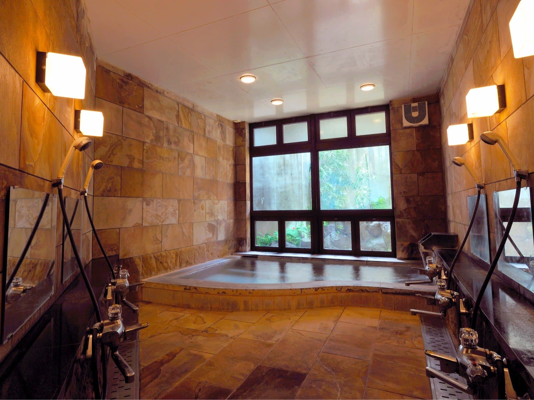 Indoor onsen bath hall at Shirouma-so with sandstone walls, washing stations and a garden-view window in Hakuba Onsen