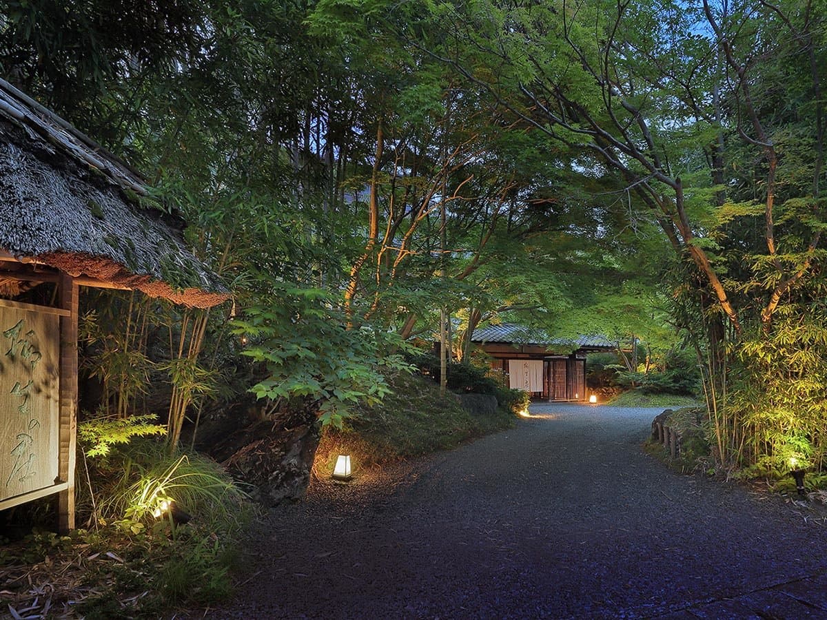 Lantern-lit gravel entrance approach to the thatched-roof main gate of Yagyu no Sho ryokan at dusk in Shuzenji Onsen, Izu