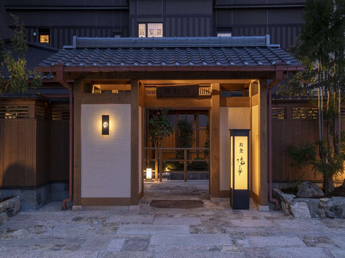 Illuminated wooden entrance gate of Noto Kaishu ryokan at dusk in Wakura Onsen, with stone path and lanterns