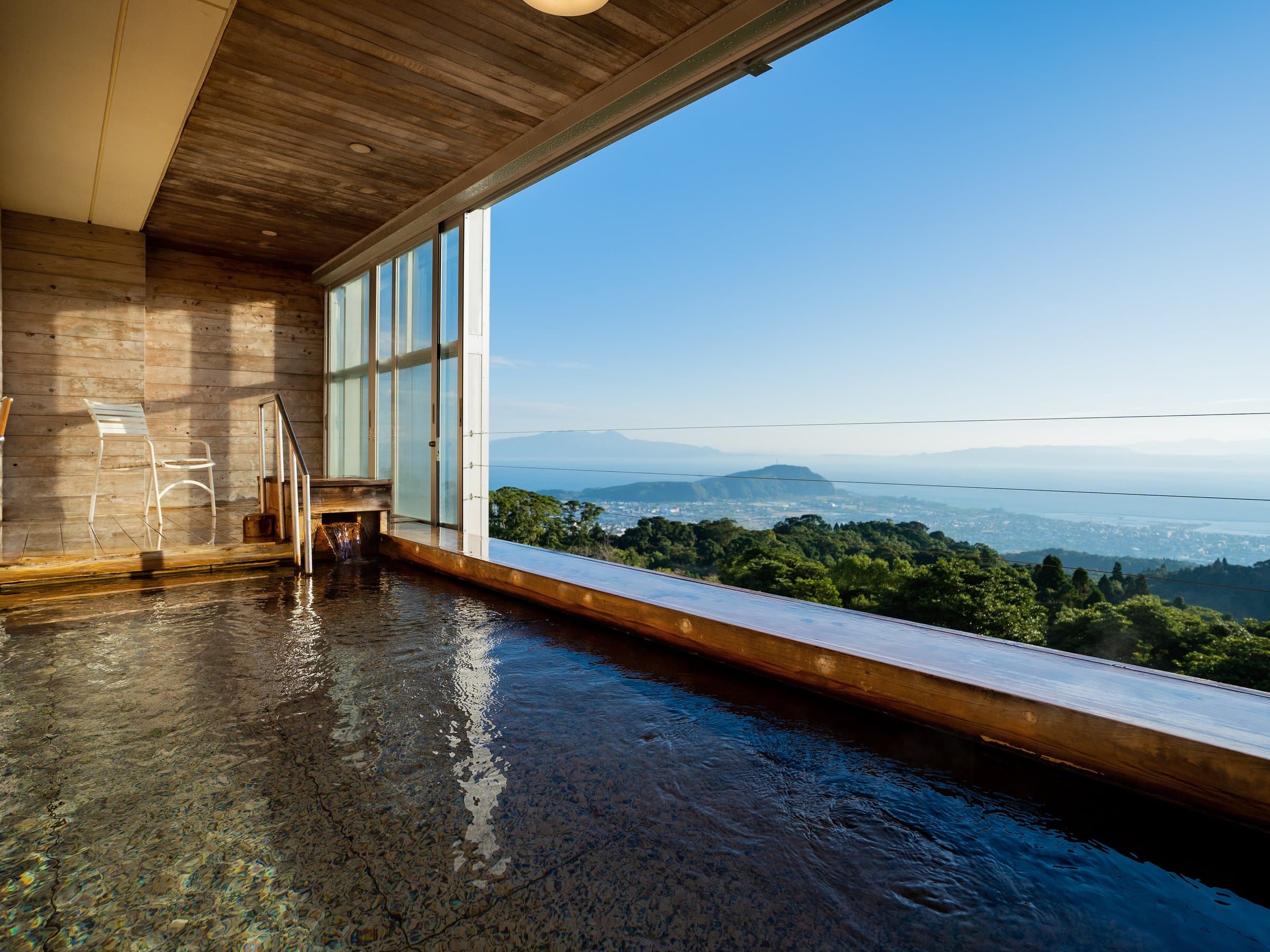 Indoor hot-spring bath with wide windows opening to a sweeping Ibusuki townscape and sea view