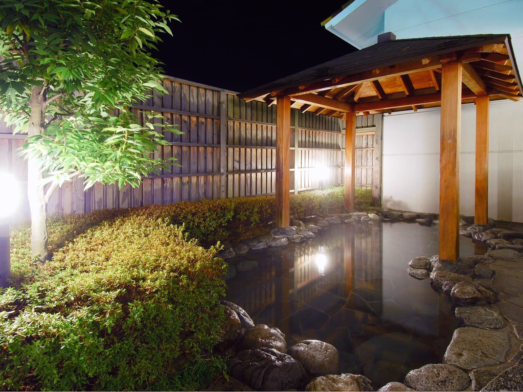 Illuminated outdoor rotenburo hot-spring bath with a wooden pavilion and garden at night at Sanshi Suimei, Izu