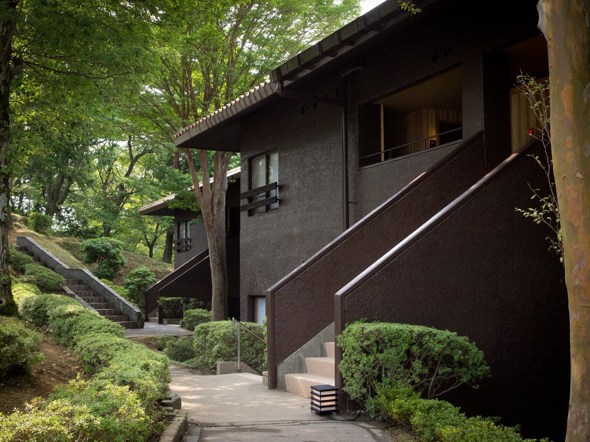Dark-walled villa wing with tiled eaves and a landscaped stone-path approach through trees at Sanshi Suimei in Shuzenji Onsen