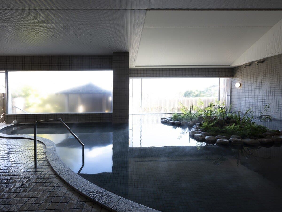Indoor public onsen bath with a rock-and-fern planted island and large windows at Sanshi Suimei in Shuzenji Onsen