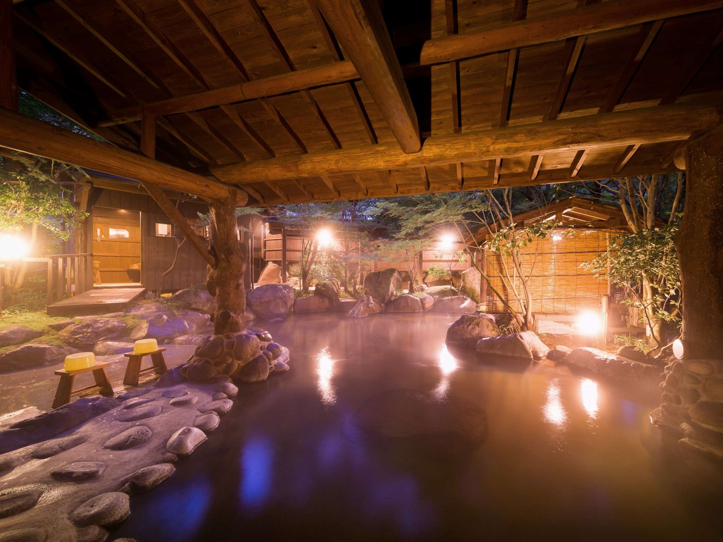 Open-air rotenburo bath at Satonoyu Waraku under a wooden roof at night, steam rising over stone-lined hot spring water