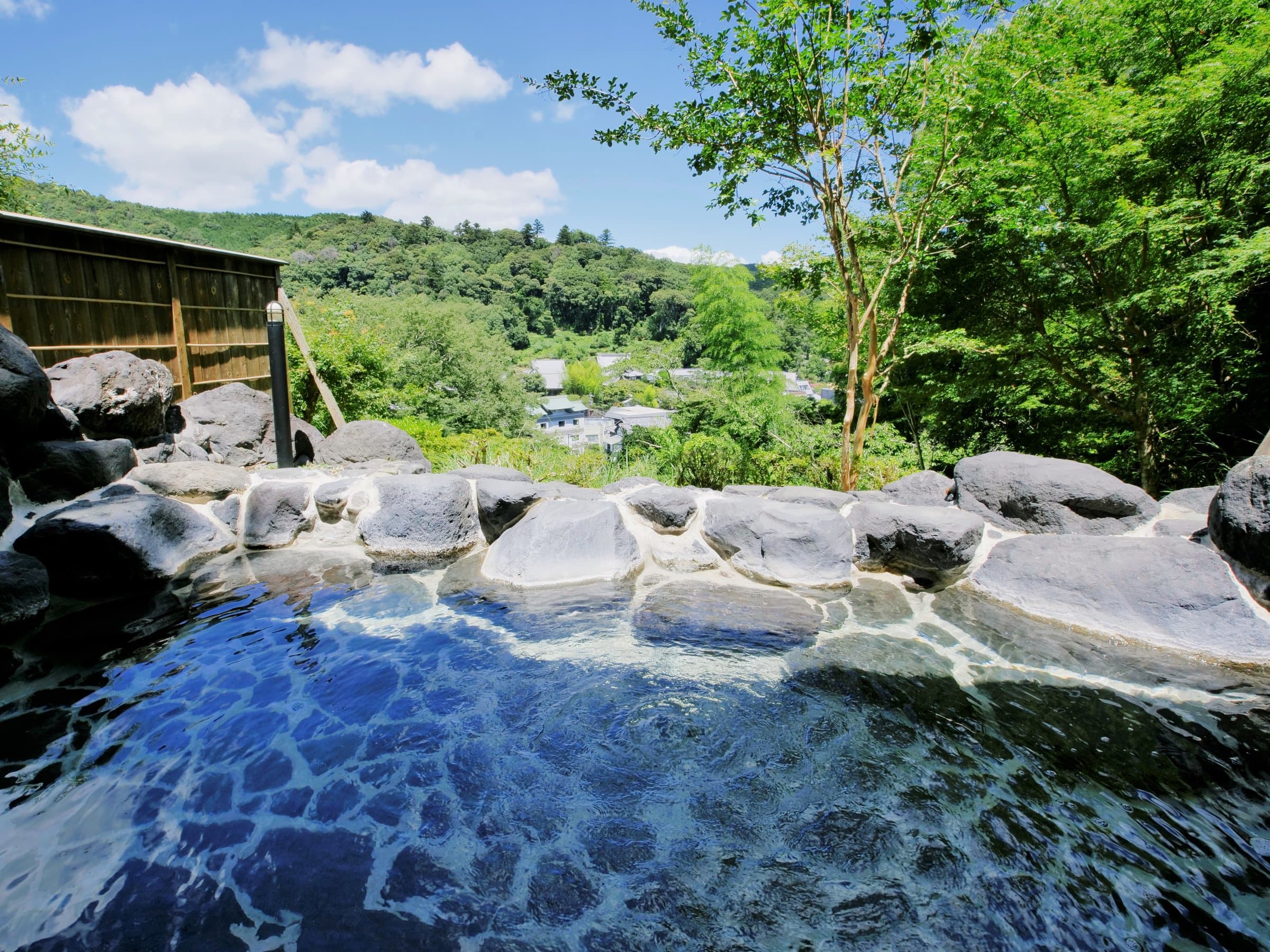 Stone-rimmed open-air rotenburo hot spring bath with a hillside town and forest view at SORA Togetsuso Kinryu, Shuzenji Onsen