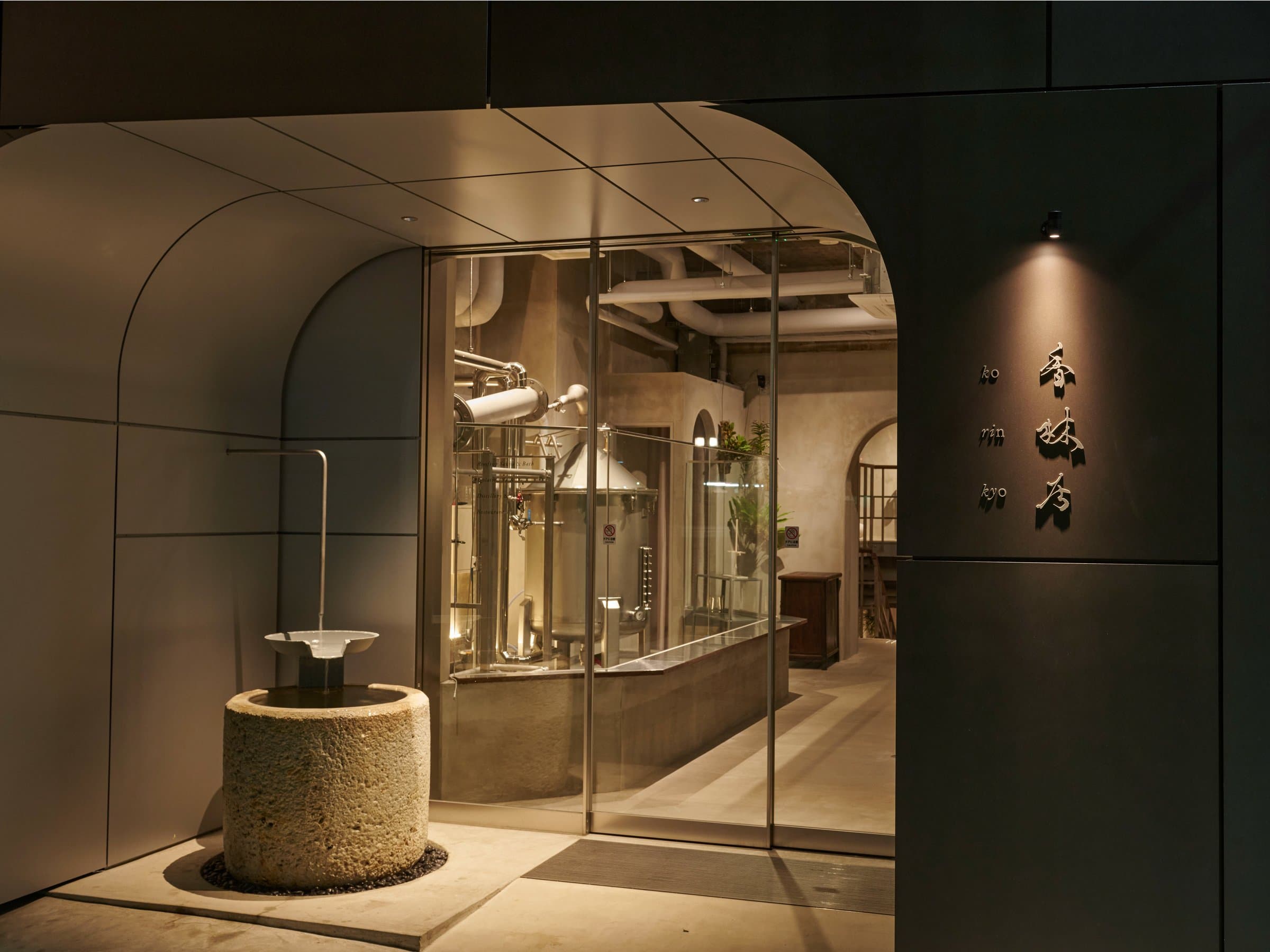 Korinkyo Kanazawa entrance at night with a stone water basin and glass-walled craft gin distillery beside the signage