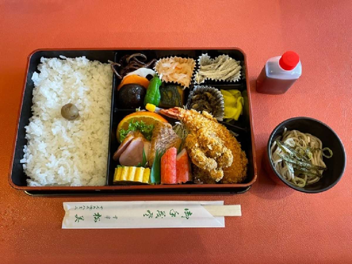 Boxed Japanese meal set with rice, fried shrimp, simmered vegetables and a small bowl of soba