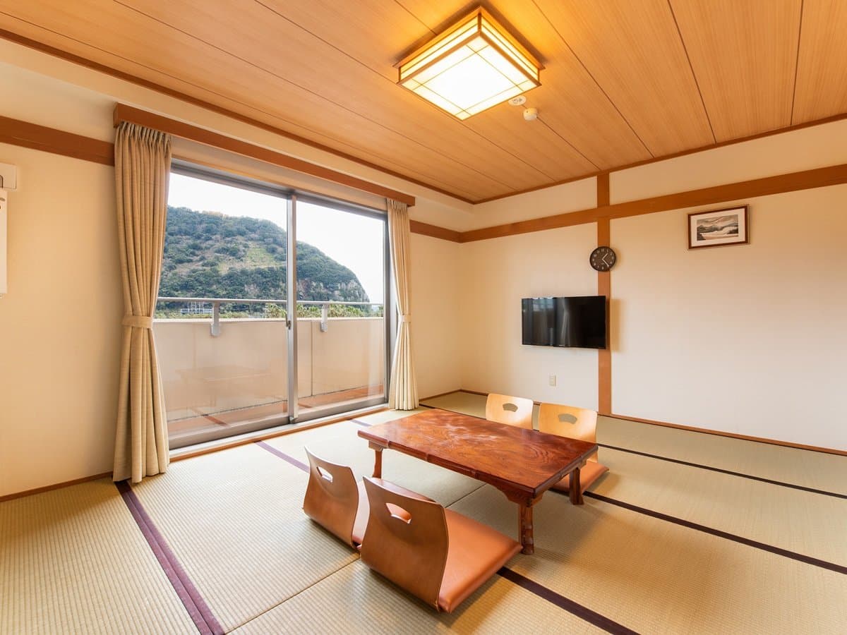 Japanese-style tatami guest room with a low wooden table, floor seating and a wide window framing a green hillside view at Ibusuki Kokoro no Yado