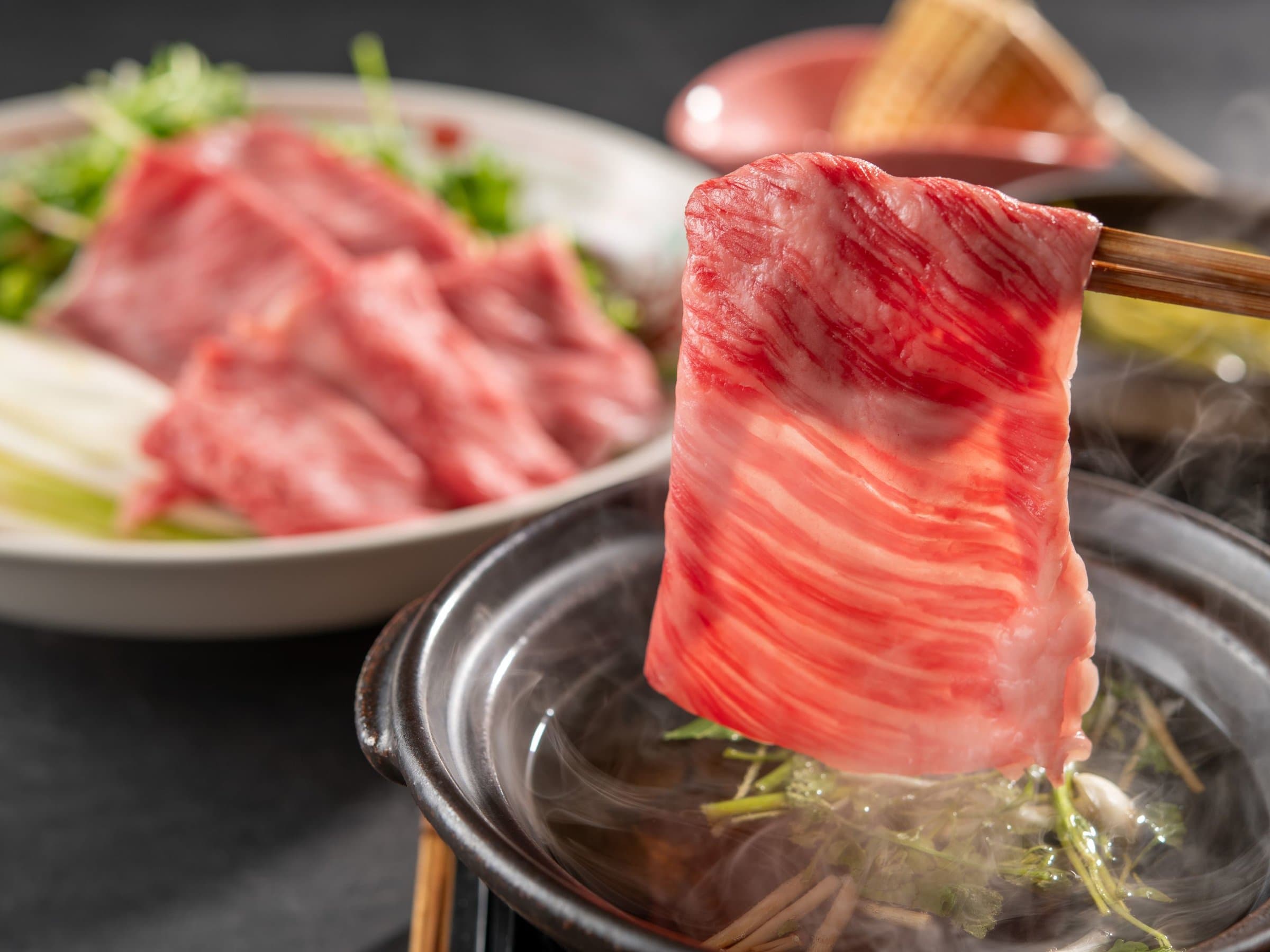 Marbled wagyu beef slice lifted over a steaming shabu-shabu hot pot with fresh vegetables