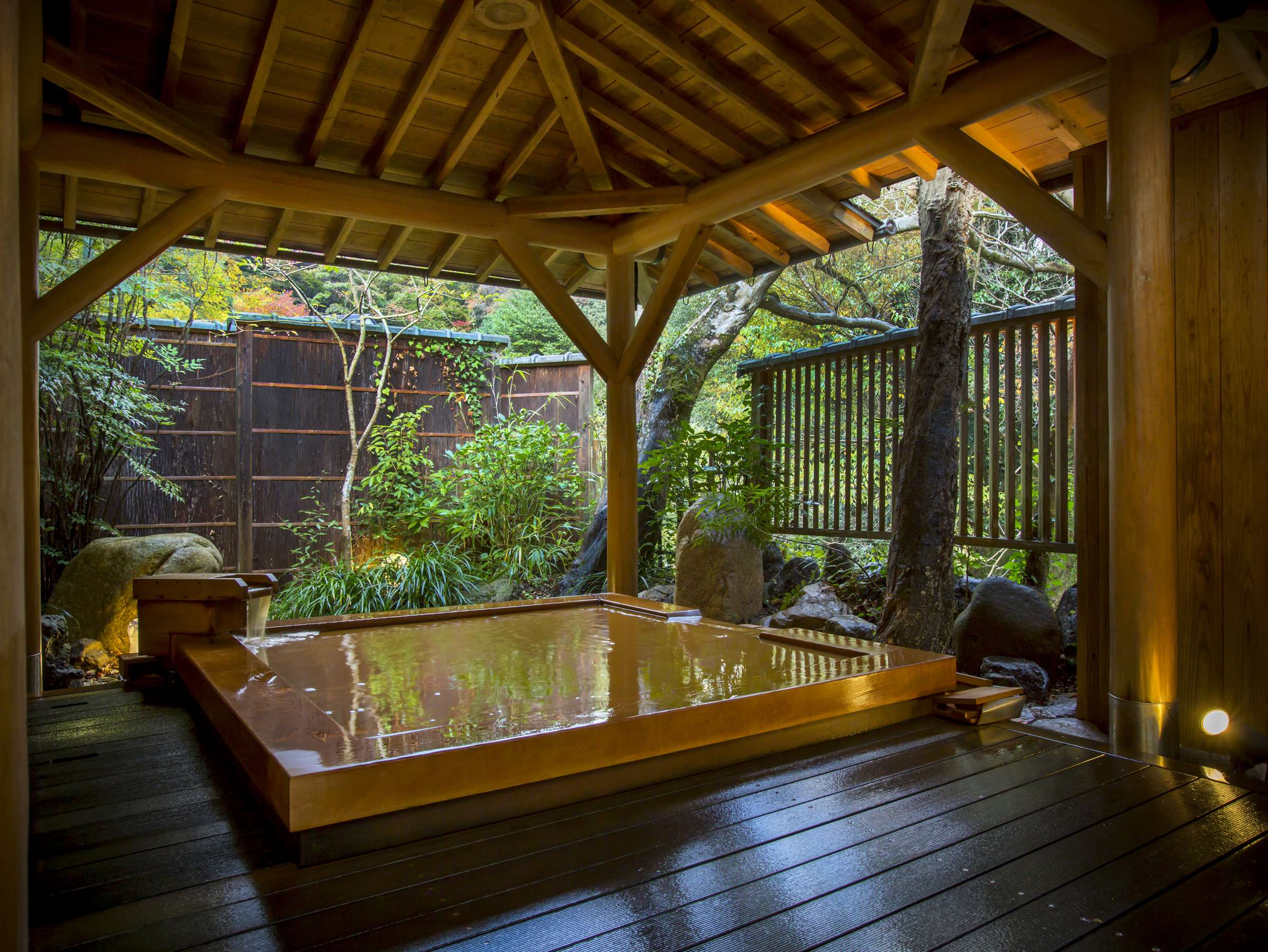 Private outdoor cypress rotenburo at Hashinoya Bekkan Ransui filled with reddish-gold kinsen water, sheltered by a wooden hipped roof and bamboo fence beside a garden