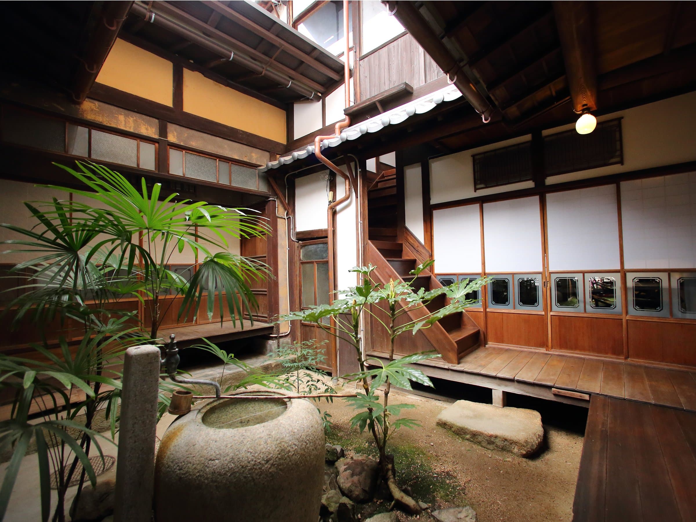 Covered tsuboniwa courtyard with stone water basin, palms and wooden staircase inside Hem's Hotel machiya, Miyajima