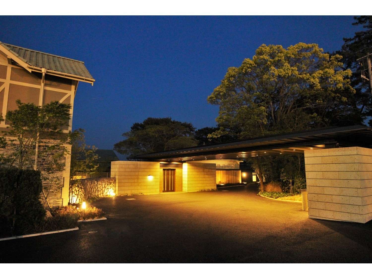 Stone entrance and covered porte-cochere lit warmly at dusk against deep blue sky and trees, Amane Resort GAHAMA, Beppu Onsen