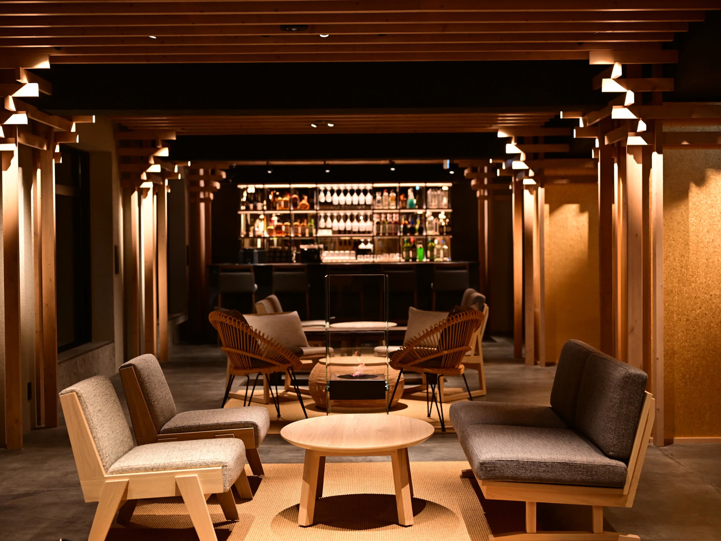 Dimly lit lounge with woven armchairs, low tables and a backlit bar surrounded by timber pillars