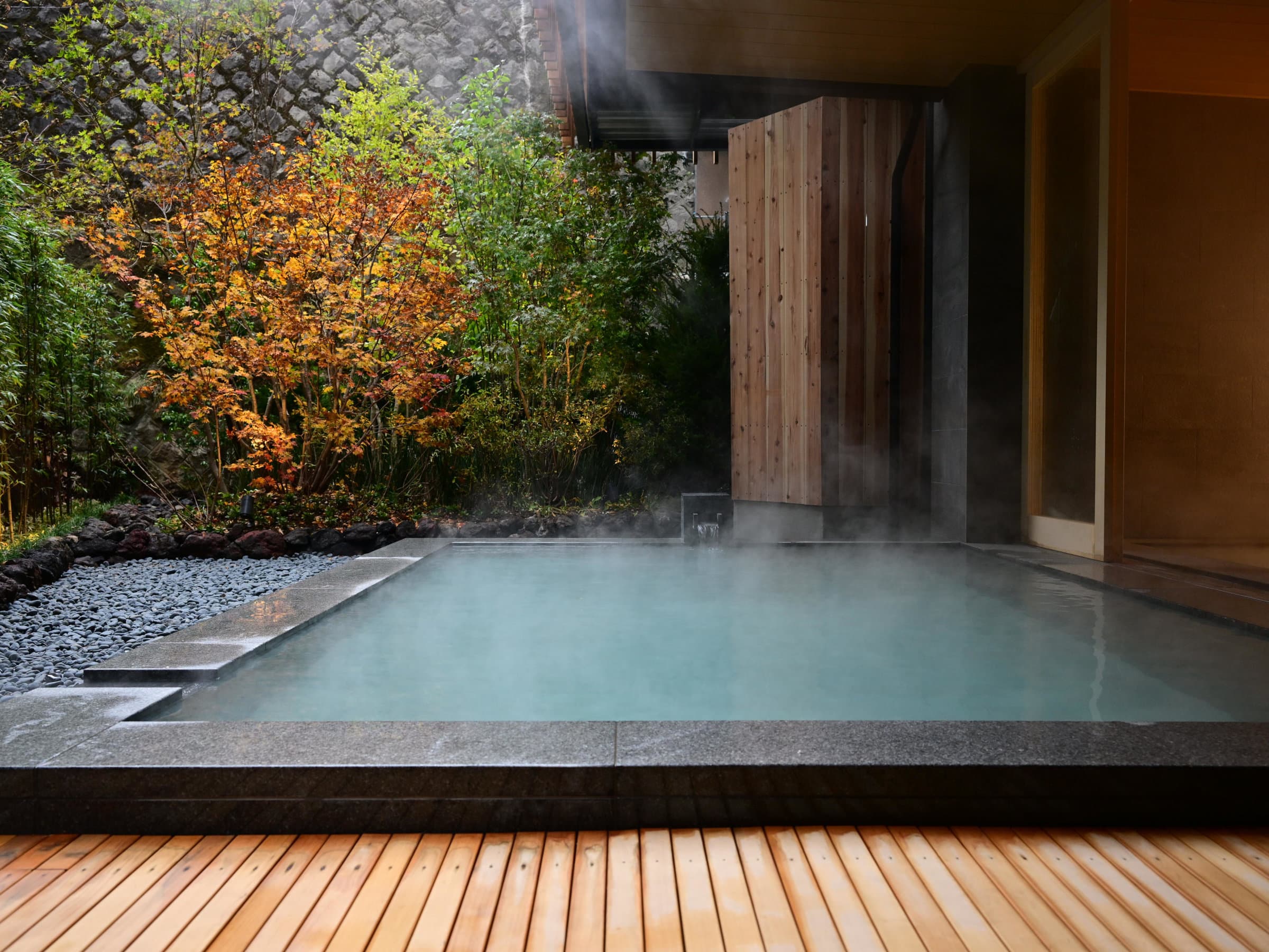 Open-air stone hot spring bath with rising steam beside autumn-tinted trees and a wooden deck