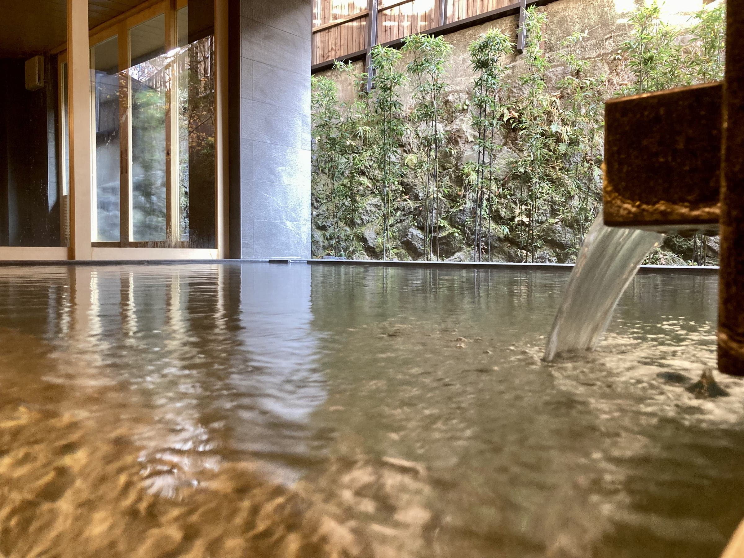 Indoor hot spring bath at water level with a rock garden and greenery seen through windows