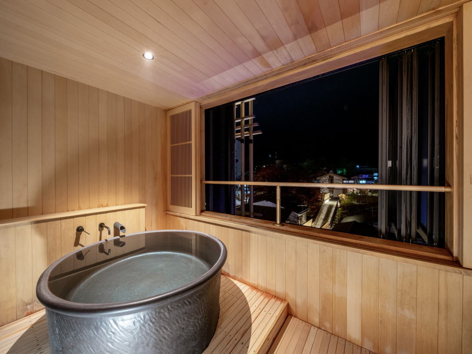 Private in-room round metal soaking tub in a cedar-lined space with a nighttime town view