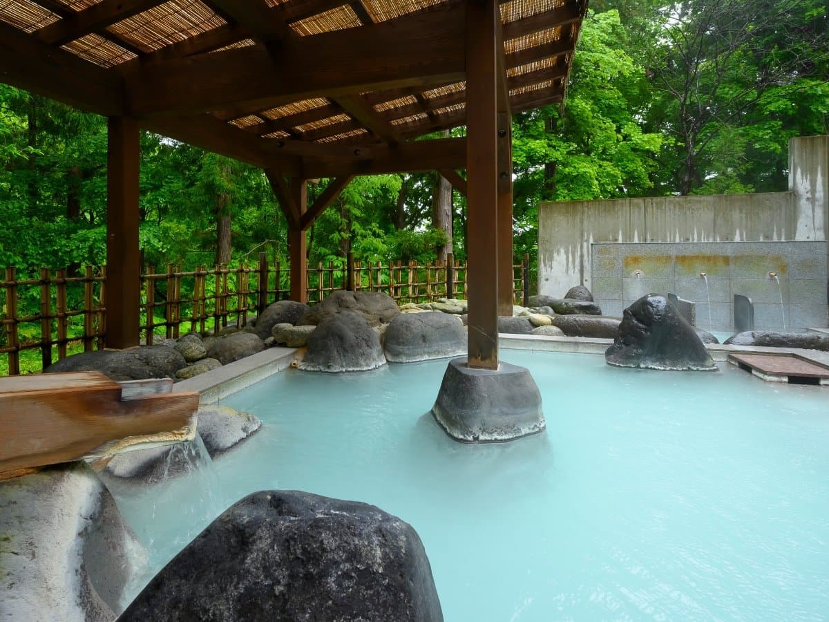 Open-air rotenburo with milky white sulfur water and rocks under a wooden roof amid forest at Zao Onsen