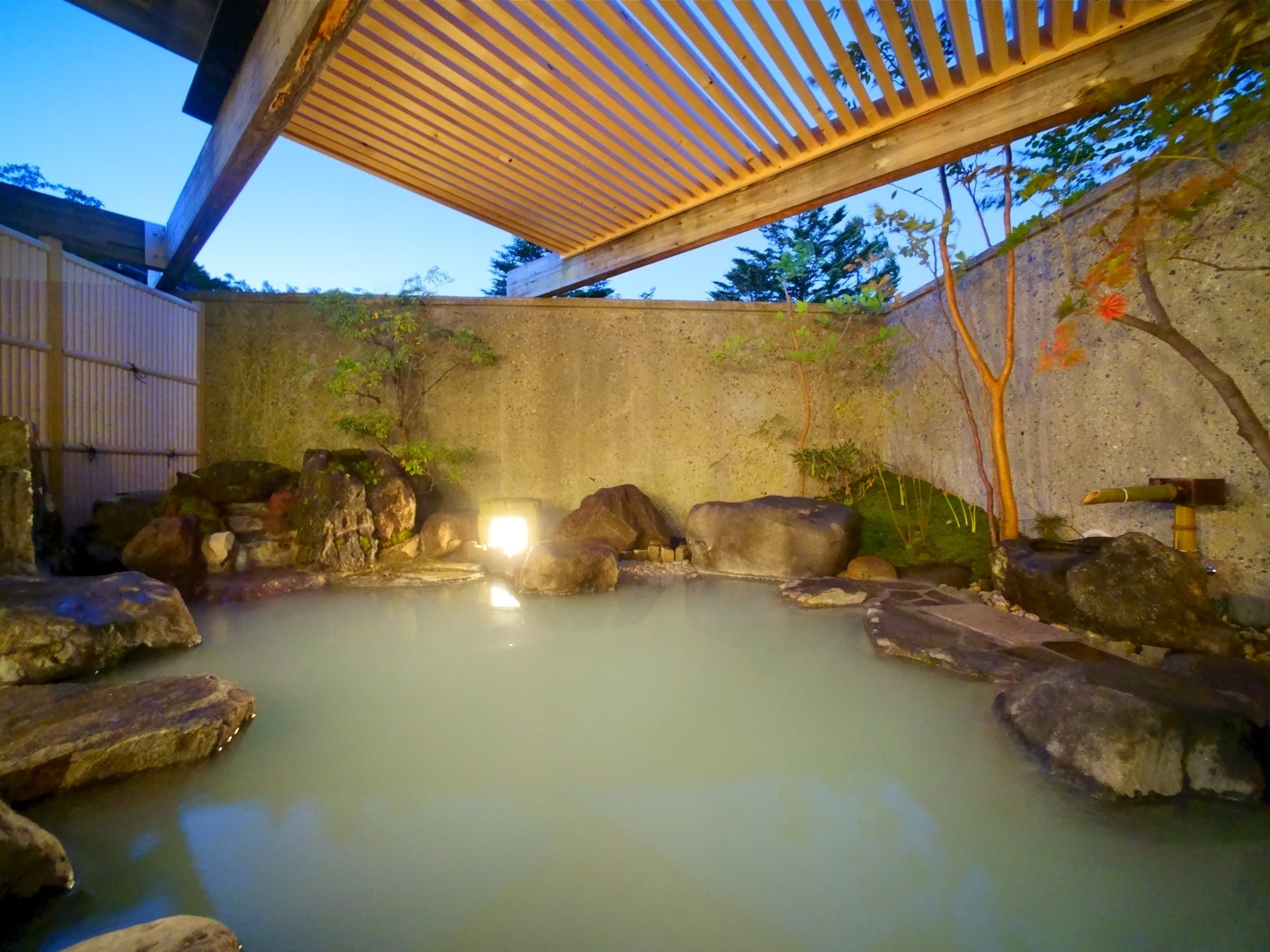 Open-air rotenburo onsen bath ringed by rocks and garden greenery at dusk at Hotel Hana-An in Okunikko