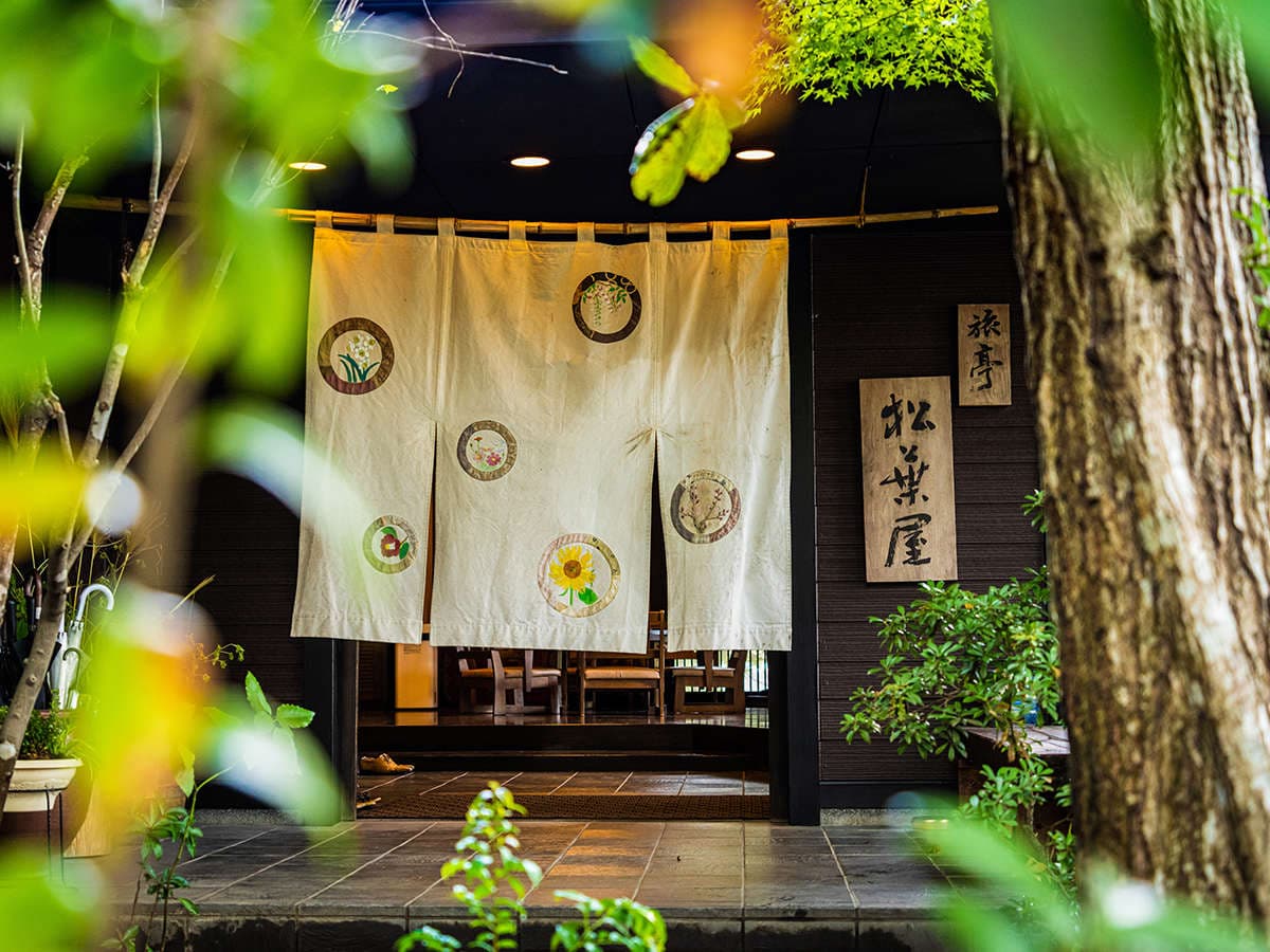 Entrance noren curtain and carved wooden inn sign glimpsed through layered green foliage at the doorway, Ryotei Matsubaya, Beppu Onsen