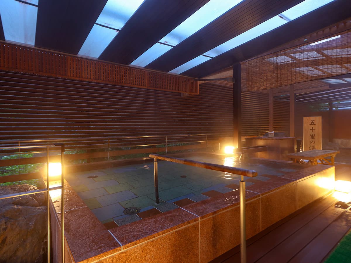 Open-air rotenburo hot spring bath under a wooden pergola lit warmly in the evening