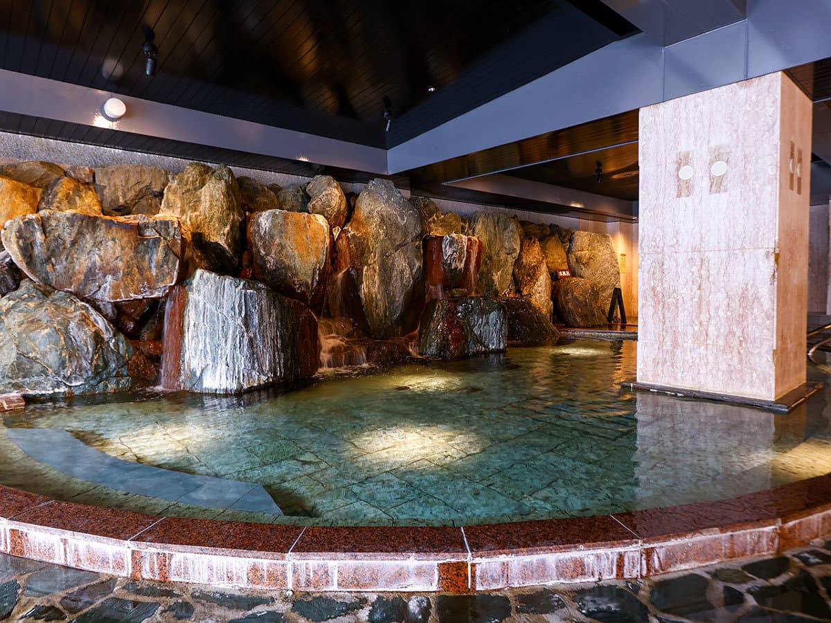 Spacious indoor stone-walled onsen bath with a cascading waterfall feature
