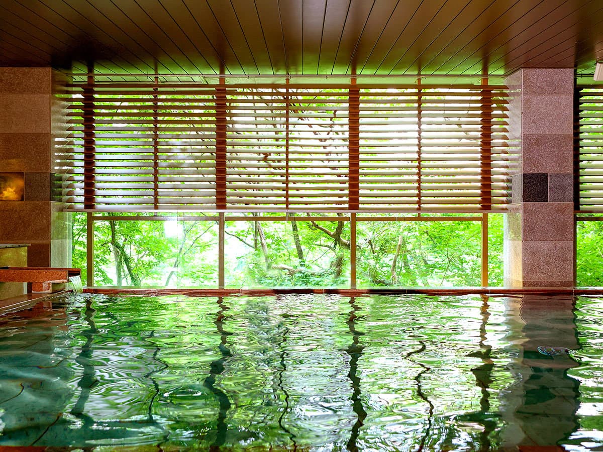 Indoor onsen bath with calm water and large windows framing fresh green foliage outside