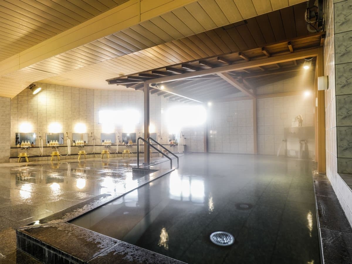 Large indoor public onsen bath with steam, stone floor and wooden ceiling at Hotel Katsuragi, Dogo Onsen