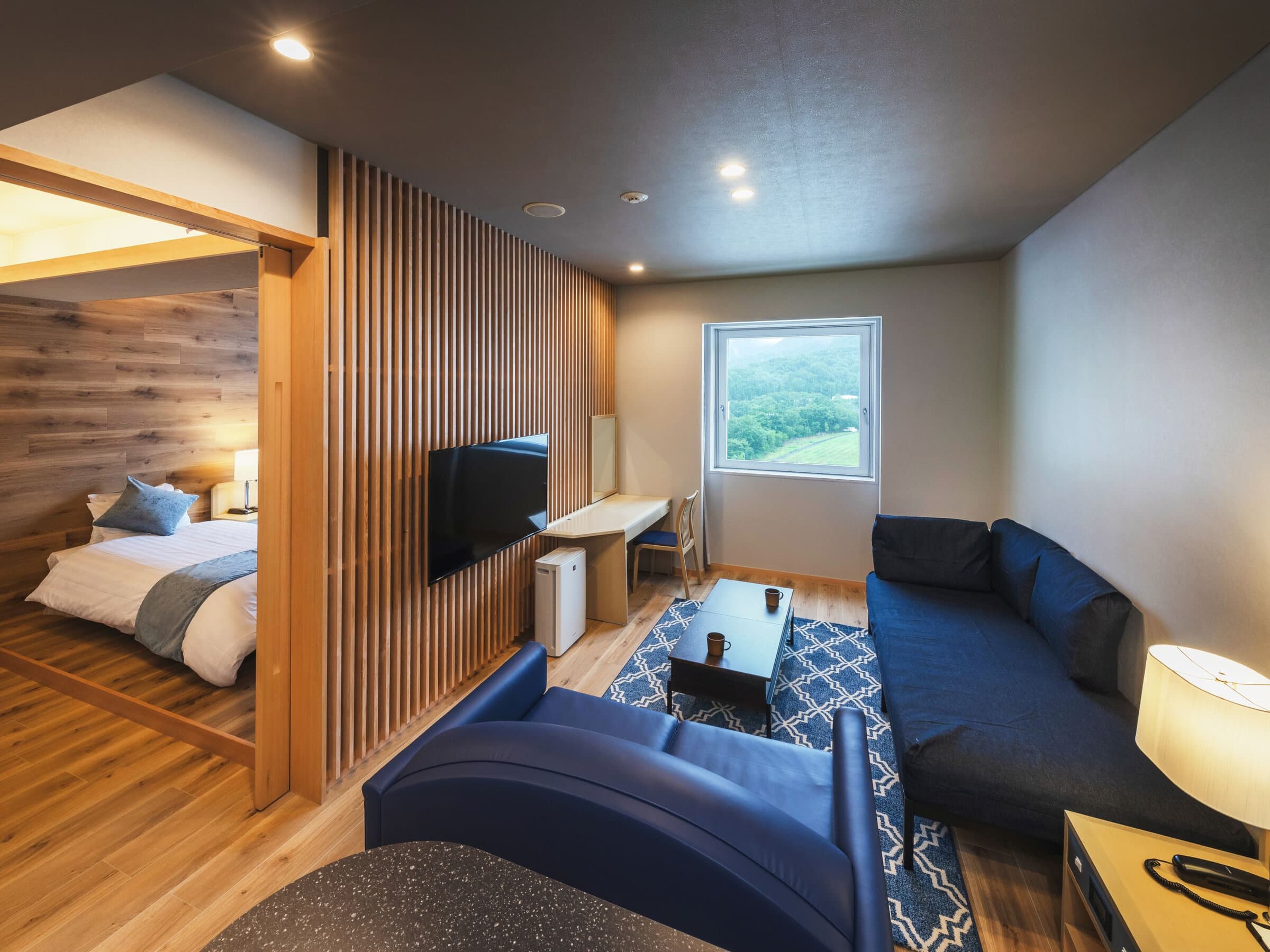 Residential suite at Takamiya Rurikura Resort with a wood-slat partition, blue sofas and a bedroom alcove