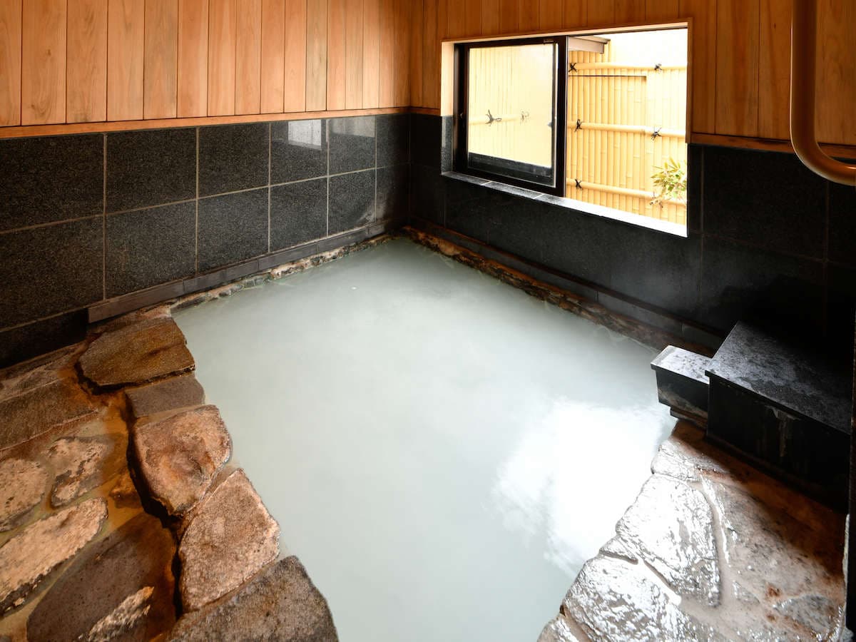 Stone-edged indoor hot-spring bath at Ryokan Tamura filled with cloudy Kusatsu spring water beside a bamboo-screened window