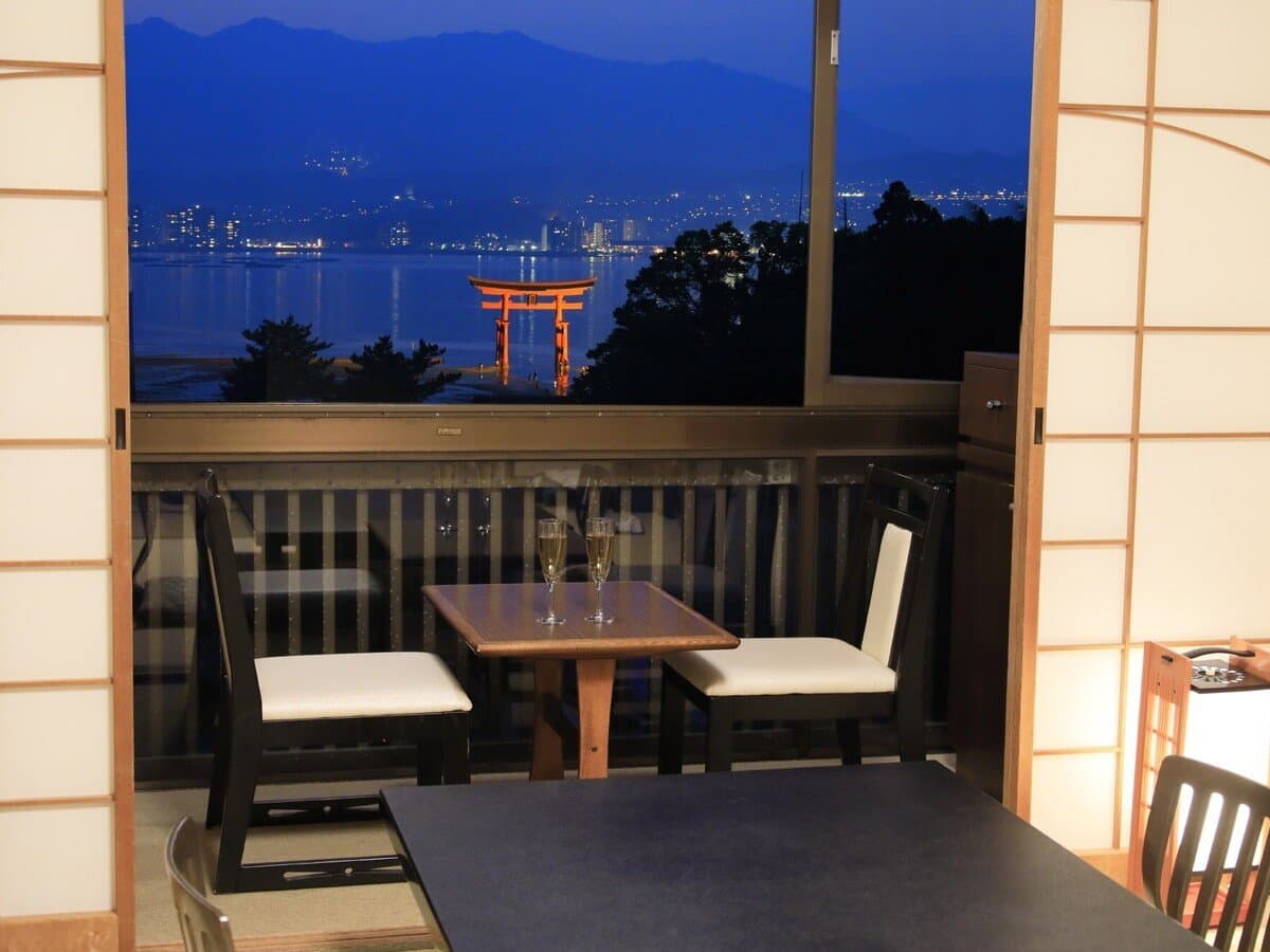 Evening dining table by a window at Hotel Kikunoya overlooking the lit floating torii gate and Miyajima coastline
