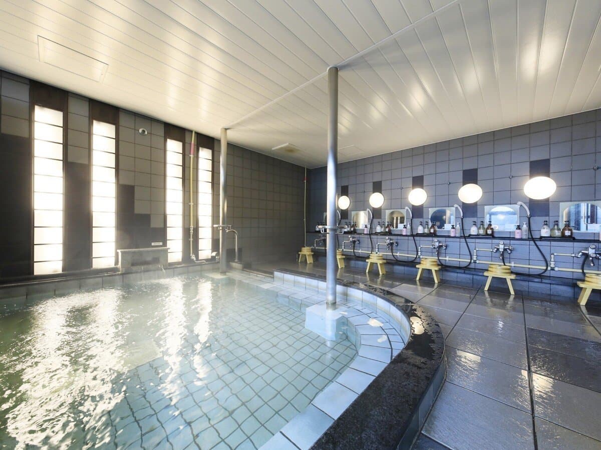 Bright indoor public bath at Hotel Kikunoya with a curved tiled tub and rows of washing stations on Miyajima island