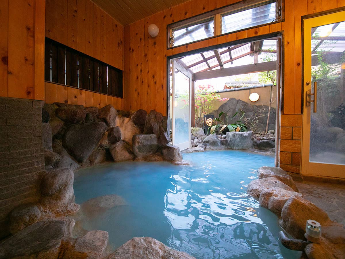 Large indoor onsen bath with natural rock surround and tall windows at Oyado Ichizen in Yufuin
