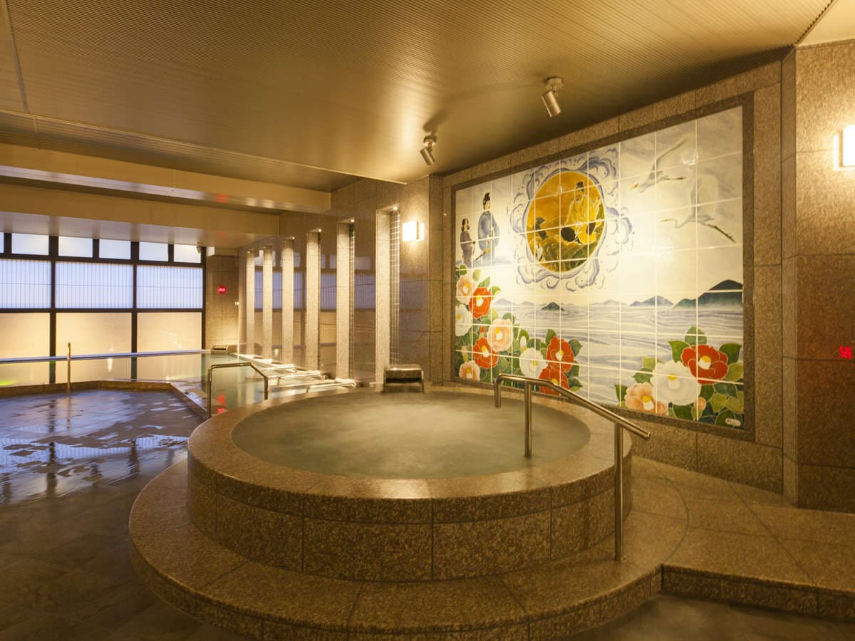 Large indoor public bath at Dogo Prince Hotel with a circular hot-spring tub and a painted Dogo Onsen tile mural