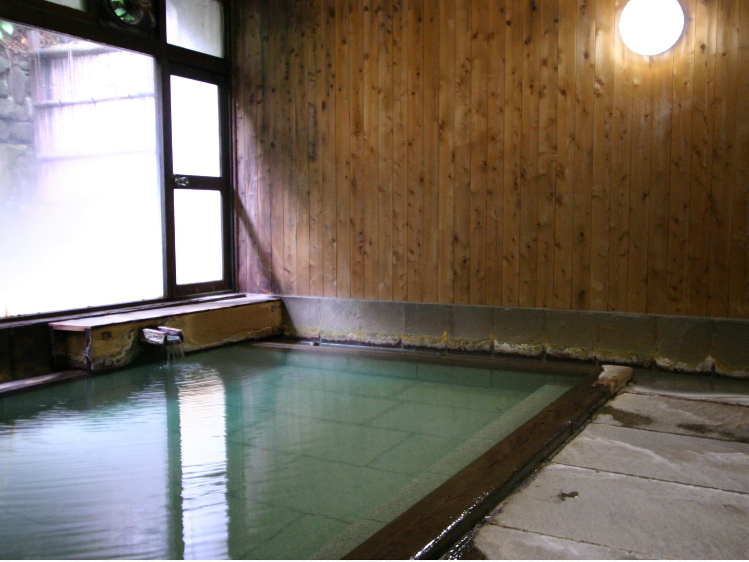 Indoor hot spring bath with wood-paneled walls and stone surround, soft natural light from a frosted window
