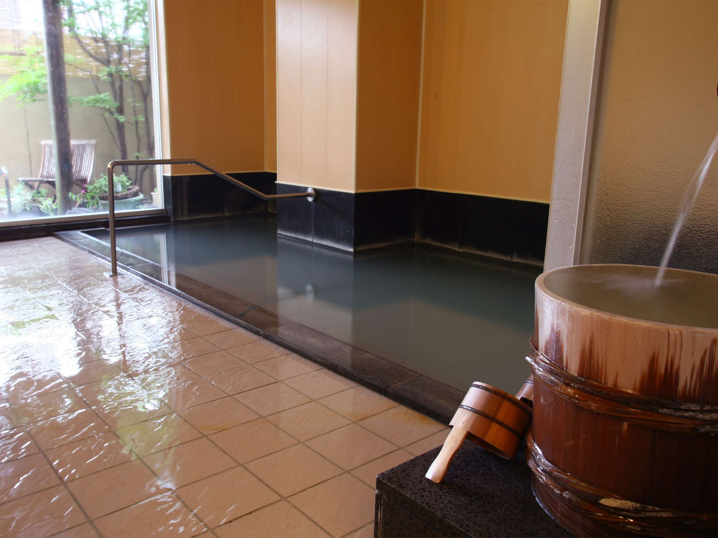 Indoor hot-spring bath with dark tiled basin and wooden ladle beside a garden window at Tamanoyu, Noboribetsu Onsen