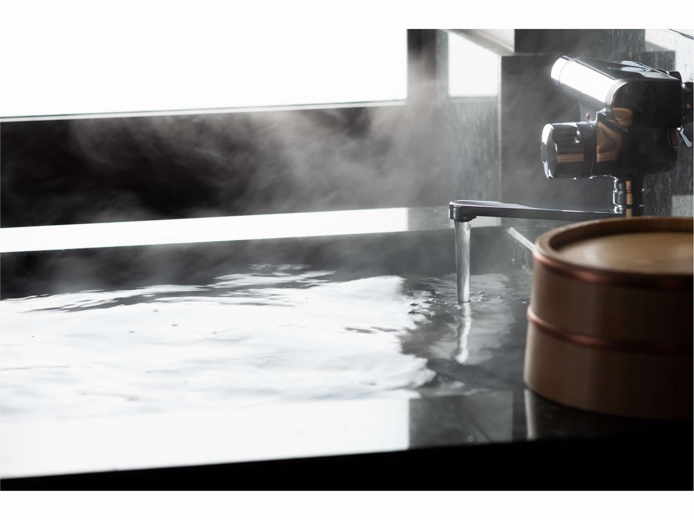 Close-up of a steaming dark-stone soaking bath with chrome spout and wooden pail at Prostyle Ryokan Asakusa