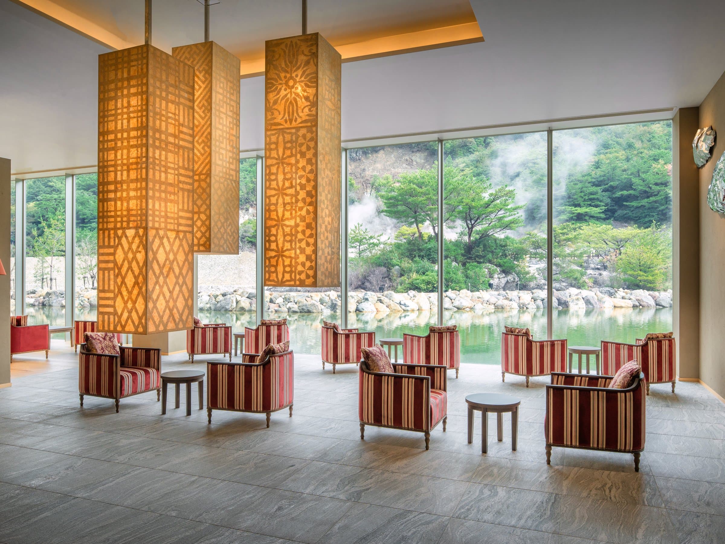 Lobby lounge at KAI Unzen with patterned lantern columns and striped armchairs before tall windows framing the steaming hot-spring landscape