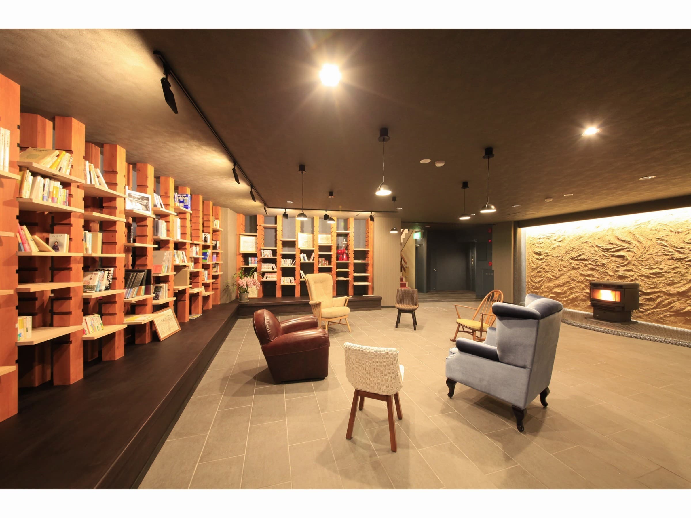 Library lounge at Yuyado Miyuki Honkan with terracotta book stacks, a wood-burning stove and armchairs on a stone floor