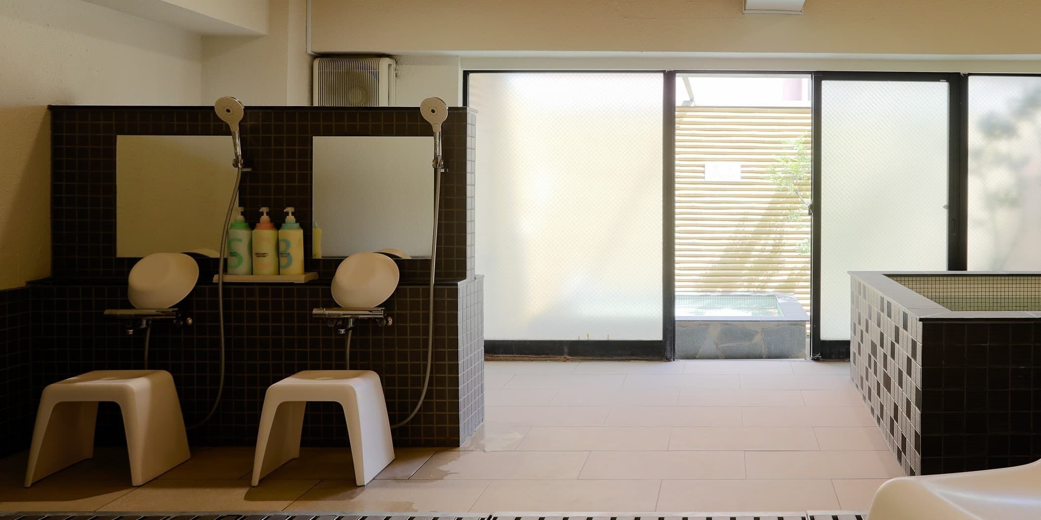 Tiled communal bath with shower stations and glass doors leading to the rooftop open-air bath at Hotel Edoya, Tokyo