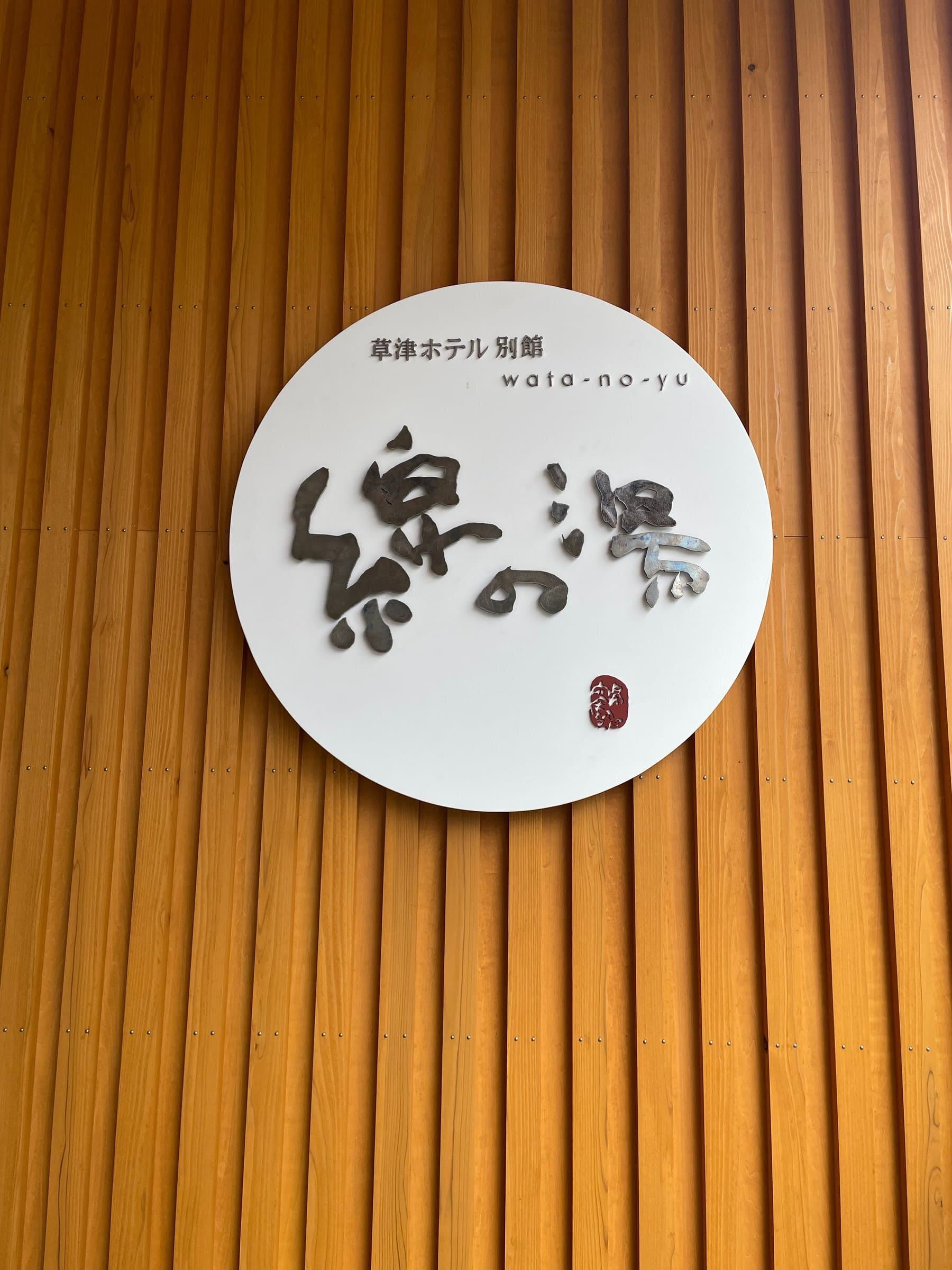 Round white entrance plaque reading Wata-no-Yu, Kusatsu Hotel Annex, mounted on a cedar-batten wall