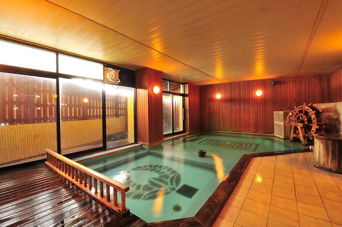 Warm wood-lined indoor onsen bath with a decorative tiled floor and stone fountain